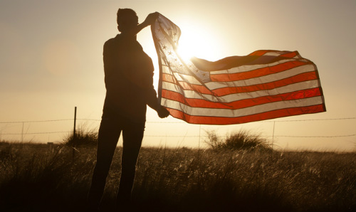 shadowed male holding american flag in front of a bright sun